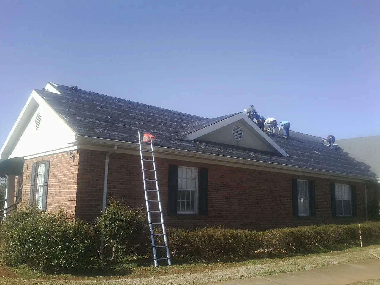 Crew tearing shingles off a brick ranch roof with a ladder set — Emergency Roof Repair work by our crew in Grand Forks Afb