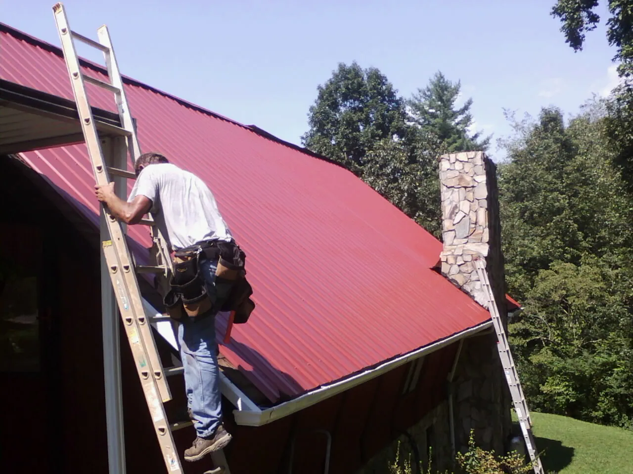Roofer on a ladder beside a red metal roof and stone chimney — Soffit & Fascia Repair by Roofing Brothers in Grand Forks Afb